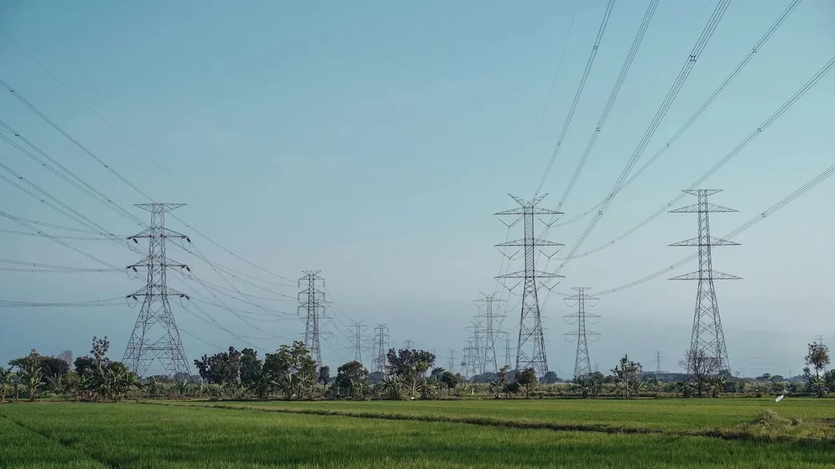 A wide landscape shot of high-voltage power lines and transmission towers stretching across a lush green field under a clear blue sky.