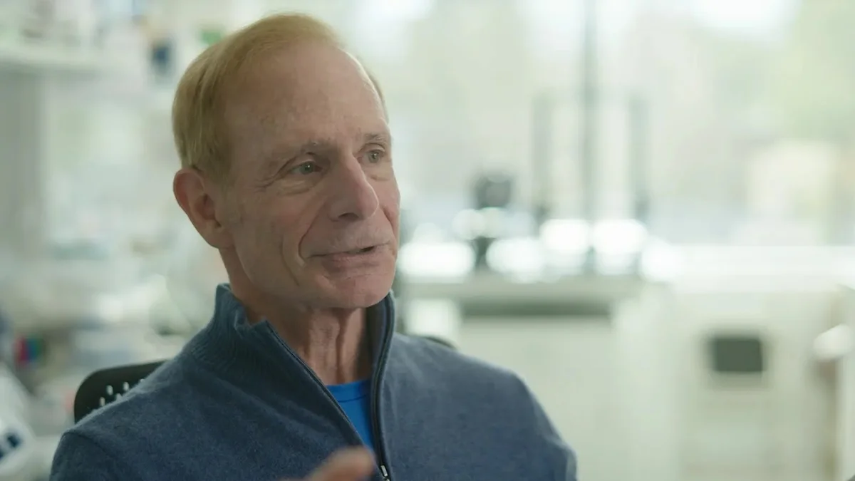 The image depicts a middle-aged man with short, thinning blonde hair, captured from the chest up. He has a fair complexion, light eyes, and is wearing a dark blue, textured zip-up sweater over a bright blue shirt. He is looking slightly off-camera toward the left, appearing to be in the middle of speaking or listening. The background is a slightly blurred laboratory or office environment with hints of equipment, suggesting a professional setting
