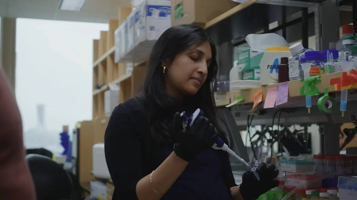 The image depicts a young woman with long dark hair, wearing a dark long-sleeved top and black nitrile gloves, working in a laboratory. She is holding a pipette and focused on a task involving lab equipment, which includes racks of tubes and other supplies visible on the shelves behind her.