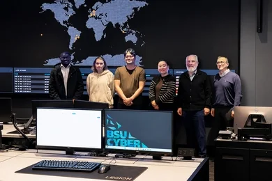 Six people pose standing in front of a wall-sized digital world map. Two computer screens dominate the foreground. One is blank and the other displays “BSU CYBER"