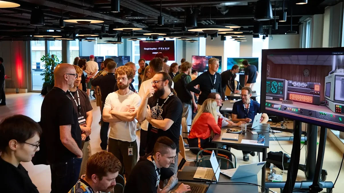 A group of people conversing and working on laptops near a large monitor showing a game demo.